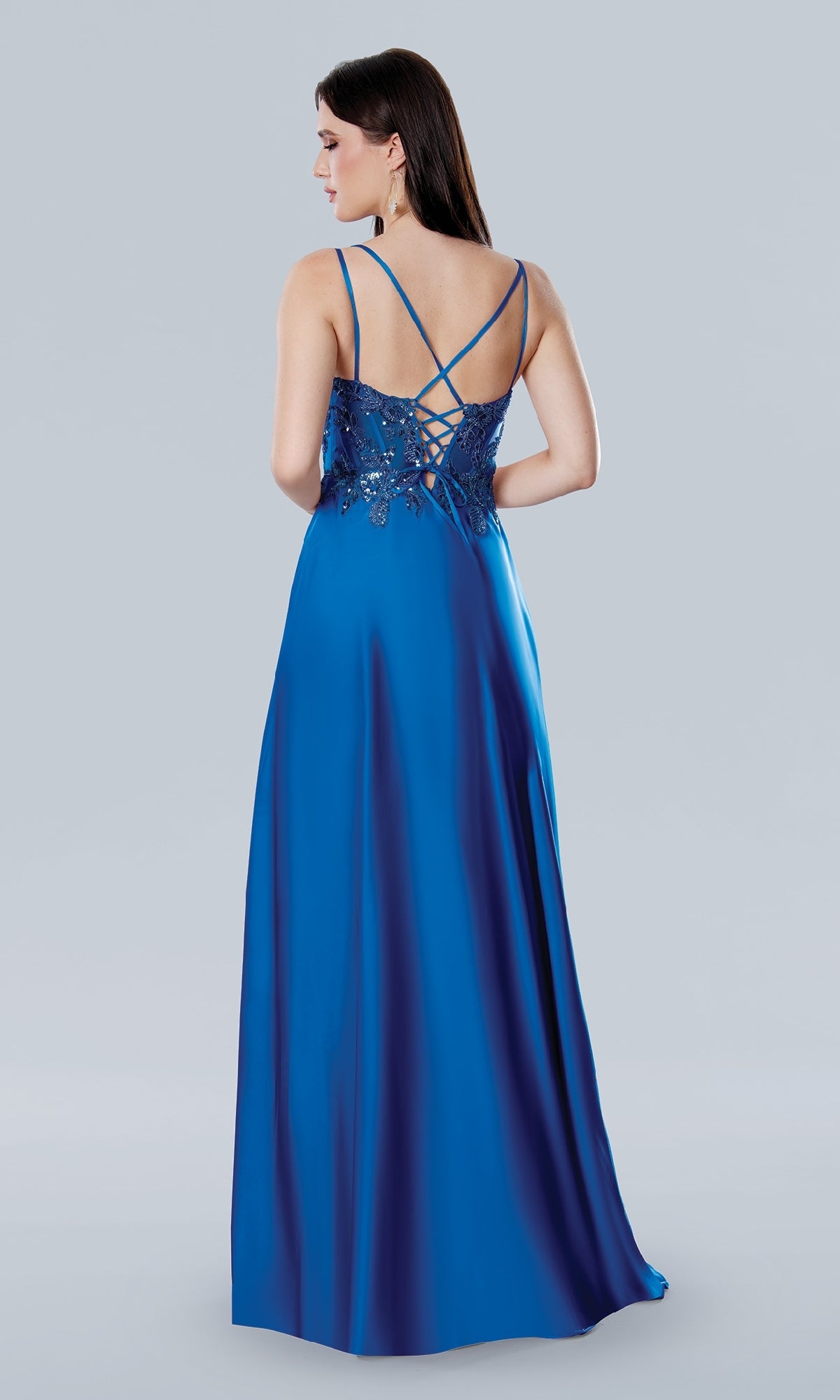A woman faces away, modeling the Stella Couture 25159 long blue satin A-line prom dress with thin crisscross straps and lace back detailing, set against a plain light gray background.
